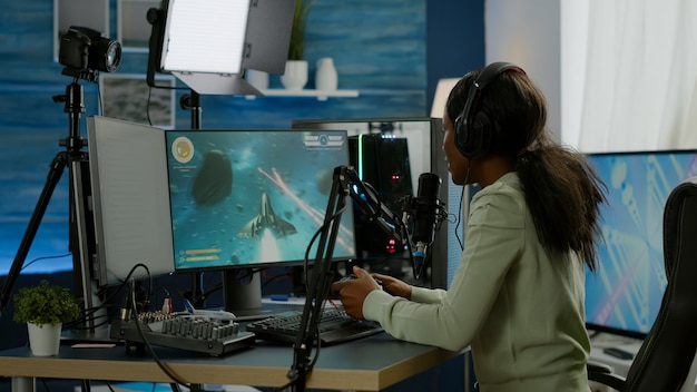 Streamer donna nera che gioca a videogiochi sparatutto spaziale con joystick che parla con i compagni di squadra in chat aperta in streaming. Cyber performance su un potente computer RGB nella sala giochi utilizzando attrezzature professionali