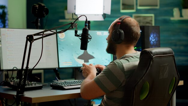 Streamer uomo che gioca ai videogiochi sparatutto spaziale utilizzando una tastiera RGB professionale e un joystick wireless. Giocatore professionista che parla al microfono durante la chat in streaming durante il torneo online