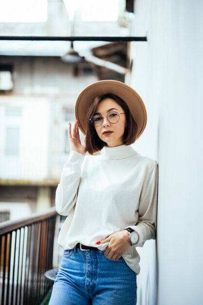 Street fashion donna interessata che indossa cappello, blue jeans, cappello largo e occhiali trasparenti sul balcone