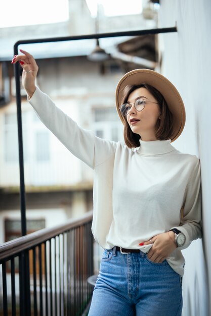 Street fashion donna interessata che indossa cappello, blue jeans, cappello largo e occhiali trasparenti sul balcone