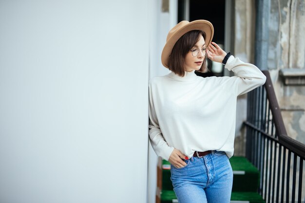 Street fashion donna interessata che indossa cappello, blue jeans, cappello largo e occhiali trasparenti sul balcone