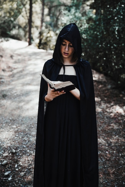 Strega incappucciata con libro in foresta durante il giorno