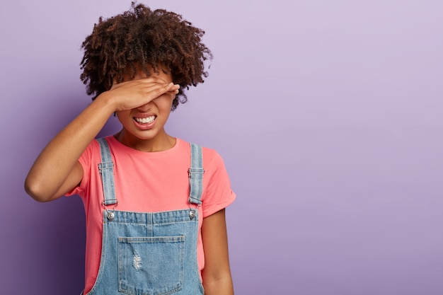 Stressante donna afroamericana copre gli occhi con la mano, stringe i denti dal dolore, soffre di mal di testa, si sente depressa, si nasconde