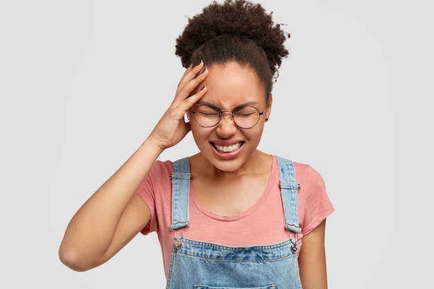 Stressante donna dalla pelle scura soffre di mal di testa, ha grossi problemi, indossa una maglietta casual e una tuta di jeans, isolata su un muro bianco. Bella giovane donna afroamericana depressa