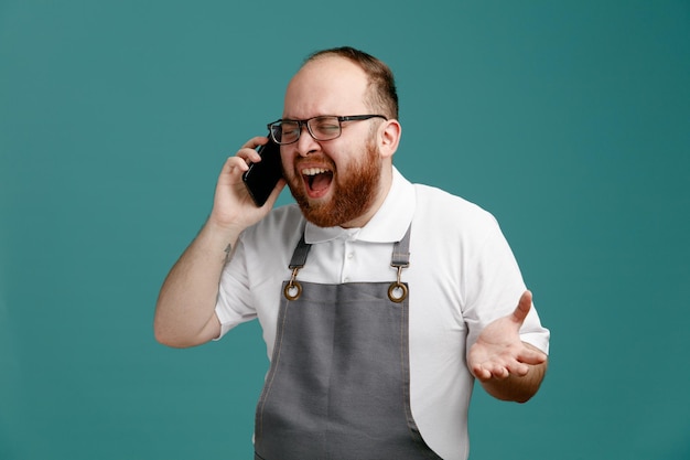 Stressato giovane barbiere che indossa uniforme e occhiali che parla al telefono gridando con gli occhi chiusi che mostrano la mano vuota isolata su sfondo blu