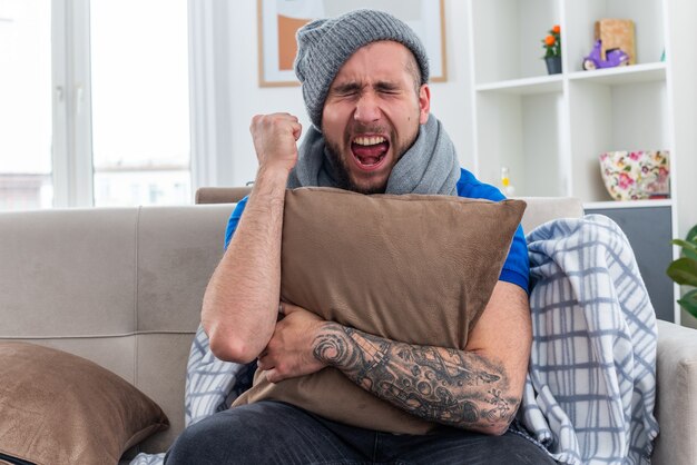 Stressato giovane uomo malato che indossa sciarpa e cappello invernale seduto sul divano in soggiorno abbracciando cuscino stringendo il pugno urlando con gli occhi chiusi