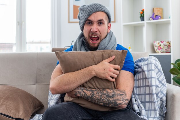 Stressato giovane uomo malato che indossa sciarpa e cappello invernale seduto sul divano in soggiorno abbracciando cuscino urlando