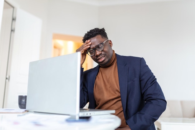 Stressato uomo afroamericano stanco che tocca le tempie che soffrono di mal di testa dopo lunghe ore di lavoro oberati di lavoro uomo d'affari sopraffatto seduto alla scrivania che si sente male