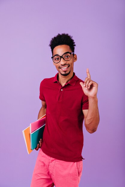 Studente africano in abbigliamento rosso in posa con un sorriso interessato. uomo nero di buon umore con gli occhiali che tiene libri ed esprime felicità.