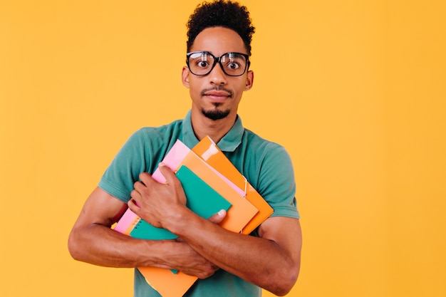 Studente africano in maglietta luminosa in posa con l'espressione del viso sorpreso. ragazzo nero con gli occhiali in piedi con i libri e alla ricerca.