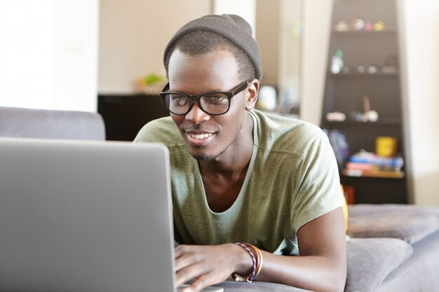 Studente afroamericano dall'aspetto alla moda che gode della connessione Internet ad alta velocità a casa, sdraiato sul divano con il notebook, guardando le serie online o giocando ai videogiochi