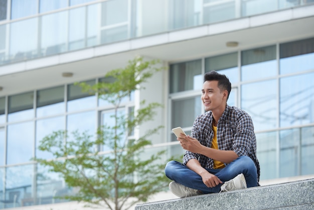 Studente asiatico che si siede sulle scale della città universitaria all'aperto con lo smartphone che fissa nella distanza
