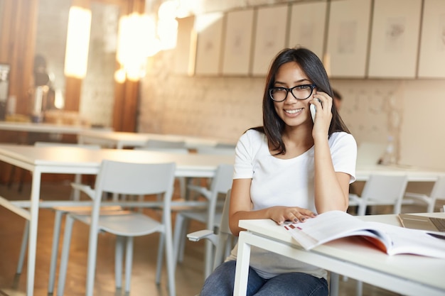 Studente che parla al telefono prendendo una pausa da lezioni o lezioni nella biblioteca openspace in chat con i suoi amici