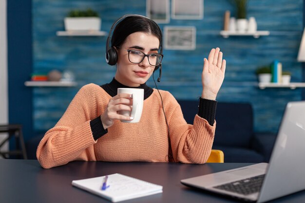 Studente che tiene una tazza di caffè mentre ascolta il suo insegnante durante la videoconferenza sul computer portatile. Donna che studia le informazioni sulla comunicazione utilizzando la piattaforma del college di e-learning