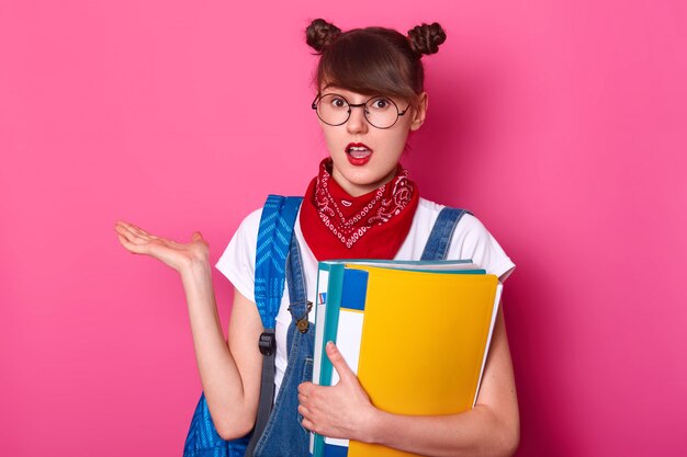 studente con due mazzi, indossa maglietta, tuta, bandana, tiene la cartella di carta, prende il palmo da parte, ha stupito l'espressione del viso, si pone con la bocca aperta isolata sul rosa.