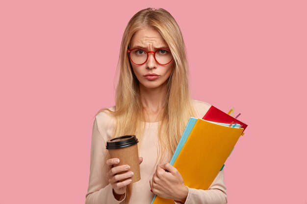 studente di college bionda insoddisfatto triste in posa contro il muro rosa