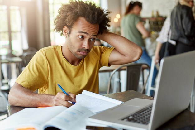 Studente di college maschio con acconciatura africana seduto alla scrivania in legno nella caffetteria scrivendo qualcosa dal computer portatile nel suo quaderno