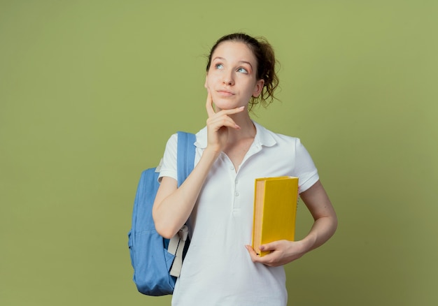 Studente femminile grazioso giovane premuroso che porta il libro della tenuta della borsa posteriore che osserva in su e che tocca il mento isolato su fondo verde con lo spazio della copia