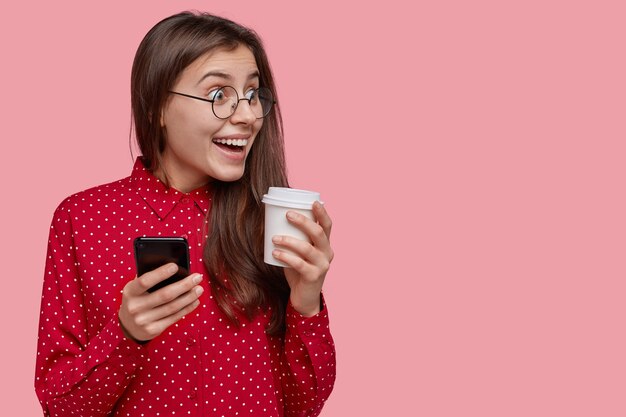 Studente gioioso con lunghi capelli neri, nota una scena divertente da parte, tiene in mano un moderno telefono cellulare e una tazza di caffè usa e getta, indossa gli occhiali