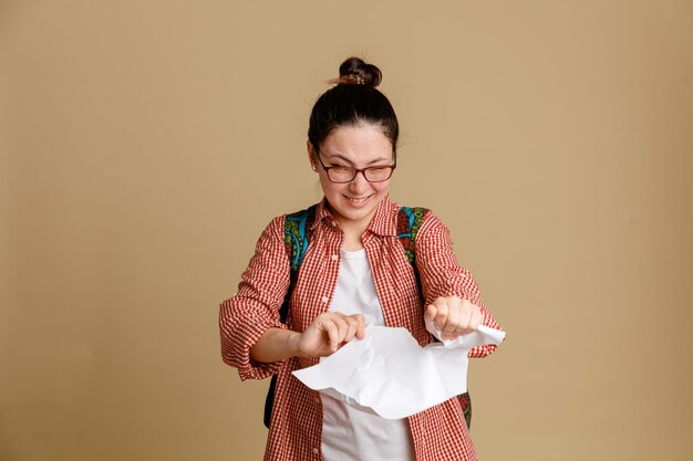 Studente giovane donna in abiti casual con occhiali con zaino strappando carta con rabbia essendo delusa in piedi su sfondo marrone
