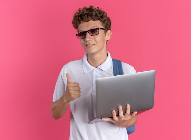 Studente in abbigliamento casual con gli occhiali con lo zaino che tiene il computer portatile che guarda la telecamera sorridendo fiducioso che mostra i pollici in su