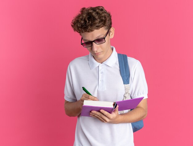 Studente in abbigliamento casual con gli occhiali con lo zaino che tiene il taccuino e la penna che scrive qualcosa con una faccia seria in piedi su sfondo rosa