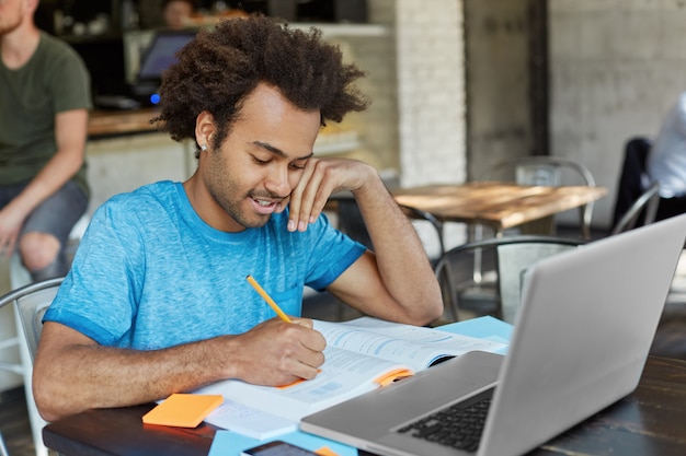 studente laureato afroamericano laborioso vestito casualmente a prendere appunti con la matita nel libro di testo mentre cercava informazioni per il documento del corso, navigava in Internet ad alta velocità sul pc portatile