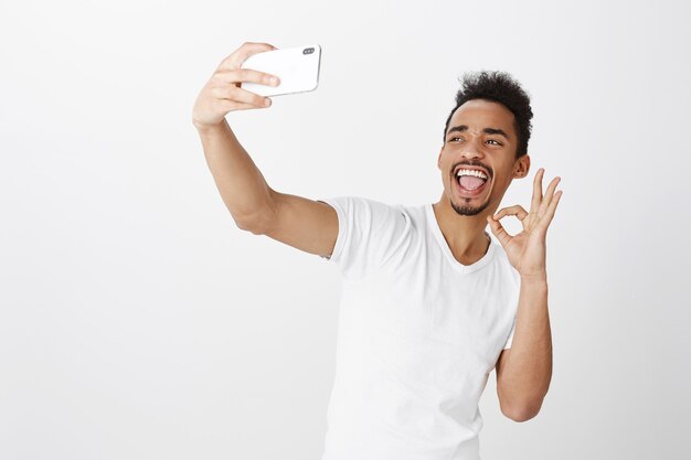 Studente maschio afroamericano sorridente impertinente che prende selfie con il telefono cellulare, mostrando il gesto giusto