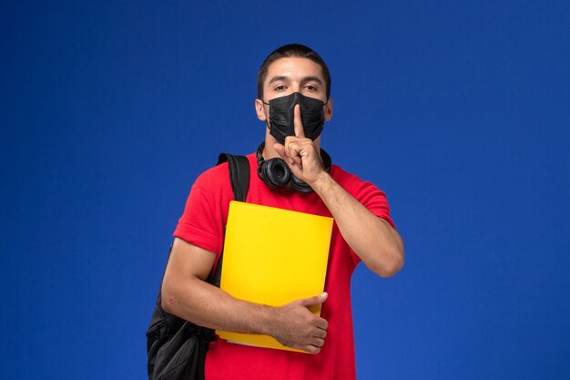 Studente maschio di vista frontale in maglietta rossa che indossa la maschera con lo zaino che tiene file giallo su sfondo blu.
