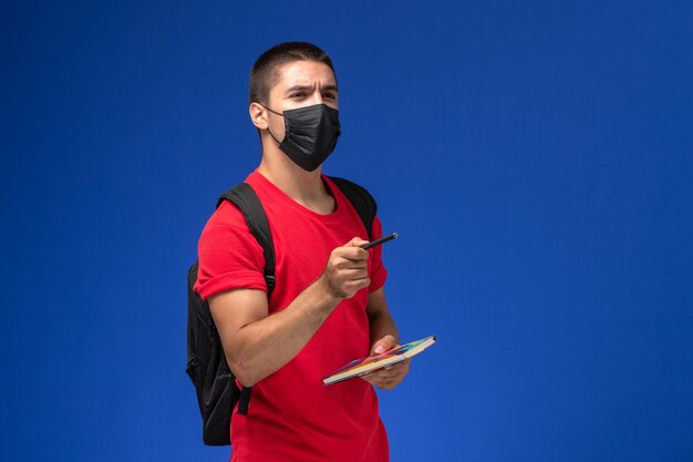 Studente maschio di vista frontale in maglietta rossa che indossa zaino in maschera sterile nera che tiene penna e quaderno sulla scrivania blu.