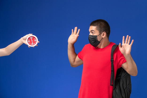 Studente maschio di vista frontale in maglietta rossa che porta zaino con la maschera spaventata degli orologi sui precedenti blu.