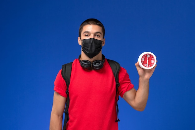 Studente maschio di vista frontale in maschera da portare della maglietta rossa con gli orologi della tenuta dello zaino sui precedenti blu.