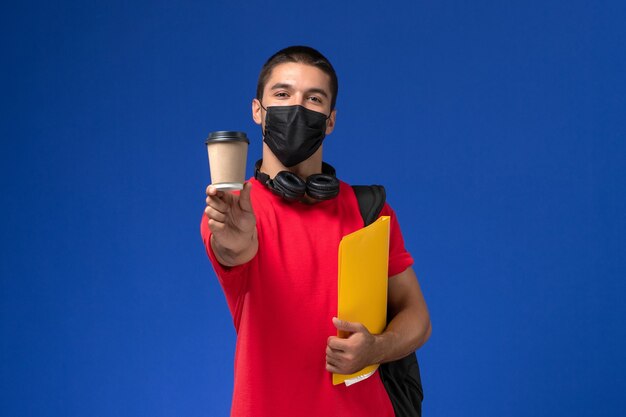 Studente maschio di vista frontale in maschera da portare della maglietta rossa con lo zaino che tiene la lima e il caffè gialli sui precedenti blu.