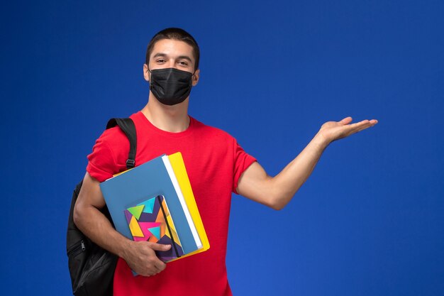 Studente maschio vista frontale in maglietta rossa che indossa zaino in maschera sterile nera che tiene quaderni su sfondo blu.