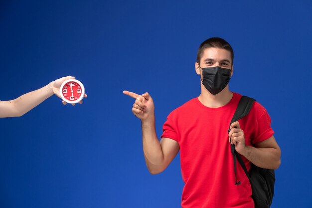 Studente maschio vista frontale in t-shirt rossa che indossa zaino con maschera in posa sullo sfondo blu.