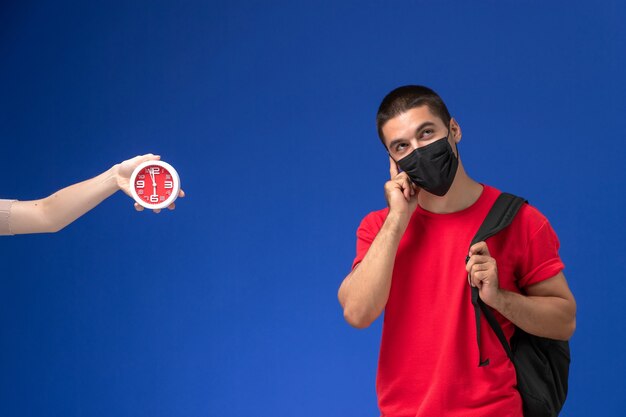 Studente maschio vista frontale in t-shirt rossa che indossa zaino con maschera pensando su sfondo blu.