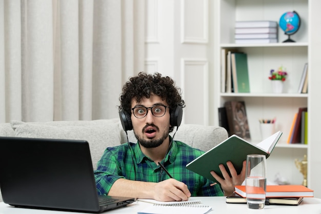 Studente online carino giovane ragazzo che studia al computer con gli occhiali in camicia verde scioccato e sorpreso