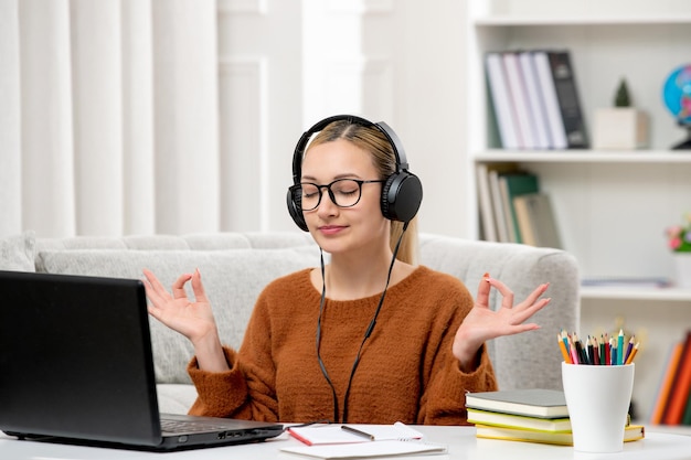 Studente online giovane ragazza carina con gli occhiali e maglione arancione che studia sul computer che mostra il segno zen