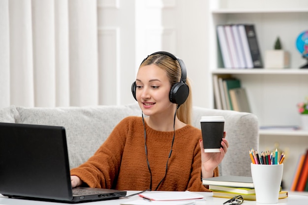 Studente online ragazza carina con gli occhiali e maglione che studia sul computer con in mano una tazza di carta