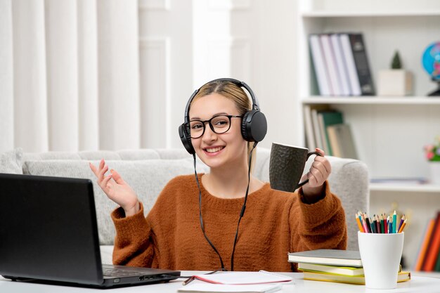 Studente online ragazza carina in occhiali e maglione che studia sul computer in possesso di una tazza di caffè