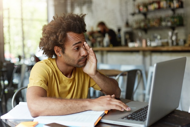 Studente universitario assonnato con i capelli folti e la pelle scura che si strofina gli occhi con la mano mentre guarda nello schermo del laptop che vuole dormire stanco preparandosi per gli esami finali.