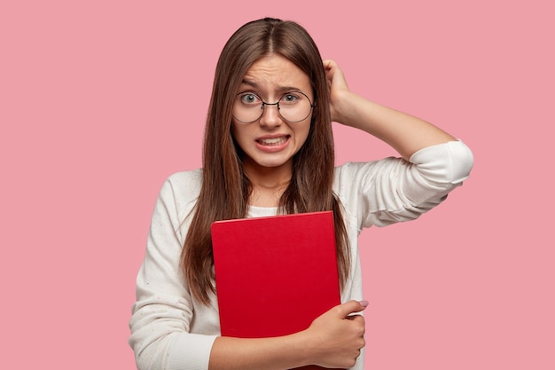 Studente universitario si gratta la testa per lo stupore, stringe i denti bianchi, ricorda le informazioni prima di rispondere all'esame finale, tiene il quaderno rosso, si sente dispiaciuto, ha problemi con lo studio.