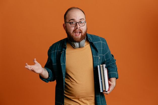 Studente uomo in abiti casual con occhiali con cuffie in possesso di quaderni guardando la fotocamera confuso e sorpreso alzando il braccio in piedi su sfondo arancione