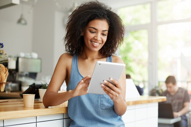 Studentessa abbastanza africana che riposa in caffè che sorride esaminando lo schermo della compressa.