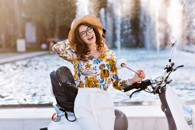 Studentessa attraente in posa giocosamente in cappello nuovo toccando il suo scooter davanti alla fontana