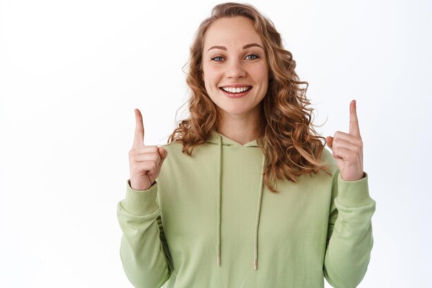 Studentessa carina allegra con capelli ricci biondi che punta le dita verso l'alto logo in alto che mostra testo promozionale pubblicitario sorridente sfondo bianco soddisfatto