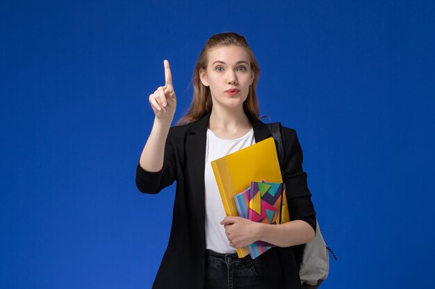 Studentessa di vista frontale in giacca nera che indossa lo zaino che tiene file con i quaderni sulla lezione dell'università dell'università della parete blu