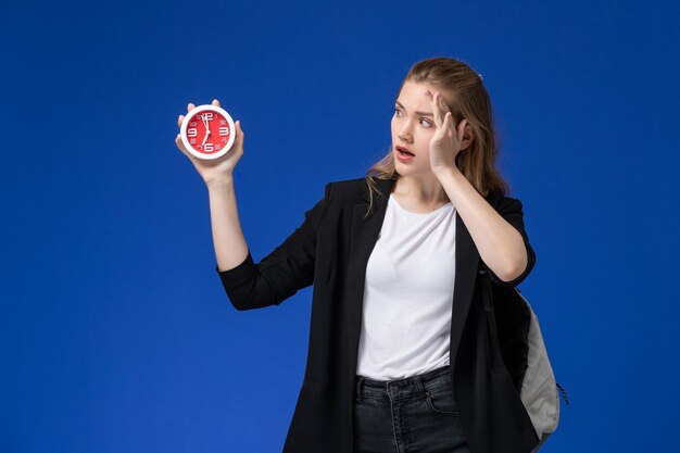 Studentessa di vista frontale in giacca nera che indossa lo zaino che tiene gli orologi sulle lezioni dell'università dell'università della parete azzurra