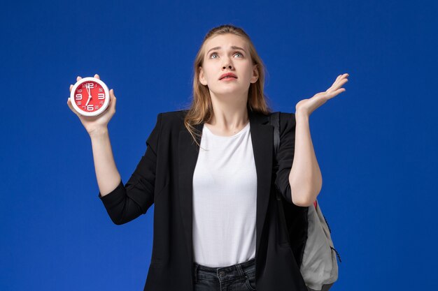 Studentessa di vista frontale in giacca nera che indossa lo zaino che tiene gli orologi sulle lezioni dell'università dell'università della parete azzurra