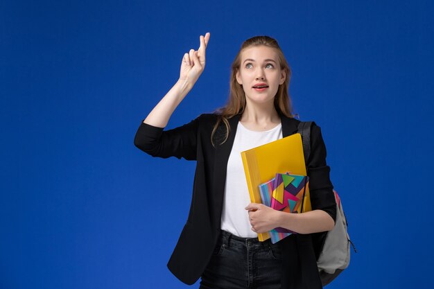 Studentessa di vista frontale in giacca nera che indossa lo zaino che tiene i file con i quaderni che incrociano le dita sulla lezione dell'università dell'università della parete blu